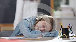 Tired Caucasian schoolgirl sleeping on desk in classroom with blurred classmate jumping at chalkboard at background