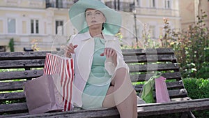 Tired Caucasian shopaholic sitting on bench on city street and sighing. Portrait of exhausted mid-adult woman with