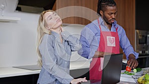 Tired Caucasian woman stretching neck spasm as blurred African American man cooking breakfast at background in kitchen