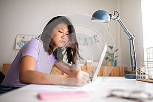 Tired chinese female college student doing homework writing on paper at home. Asian girl studying with laptop.