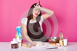 Tired cook stands with closed eyes at table in kitchen, keeps hand on forehead, spends herself with kneading dough. Young female
