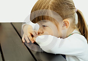 A tired cute little girl is bored sitting at a wooden table in a cafe. An angry Pretty Little kid waiting. A sad punished girl sit