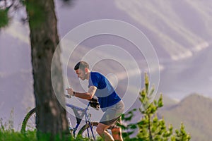 Tired cyclist is wiping his sweat off his face while pushing his bicycle uphill on a dirt road in a mountain