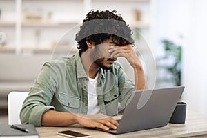 Tired dark-skinned guy sitting at workdesk in front of laptop