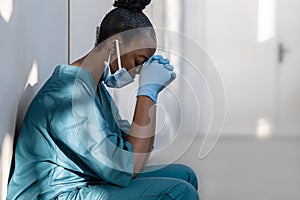 Tired depressed female african nurse wearing face mask sits on hospital floor.