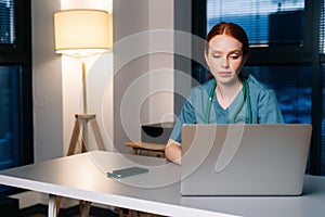 Tired depressed redhead young female doctor in blue green medical uniform working typing on laptop computer.