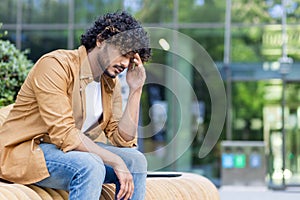 Tired and depressed young indian man sitting on a bench outside, smiling sad and holding his head