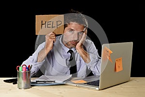 Tired desperate businessman in stress working at office computer desk holding sign asking for help