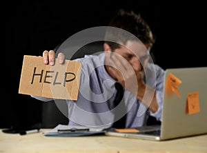Tired desperate businessman in stress working at office computer desk holding sign asking for help