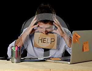 Tired desperate businessman in stress working at office computer desk holding sign asking for help