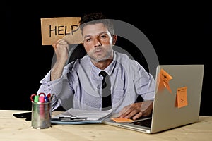 Tired desperate businessman in stress working at office computer desk holding sign asking for help