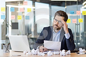 Tired disappointed bankrupt businessman sitting at workplace inside office, man in business suit depressed among papers
