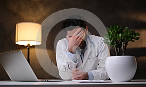 Tired doctor sitting at the table with hand at his forehead in room photo