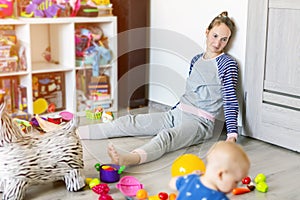 Tired of everyday household mother sitting on floor with hands on face. Kid playing in messy room. Scaterred toys and disorder. photo