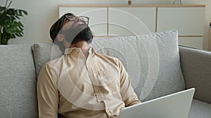 Tired exhausted freelancer working with laptop at home overworked Arabian muslim man fall asleep on couch sick weary