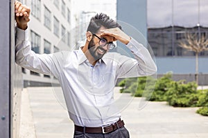 Tired and exhausted Indian young businessman standing near working offices outside and holding his hand, feeling pain