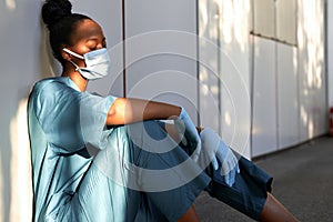 Tired exhausted woman african nurse wear face mask gloves sit on hospital floor.