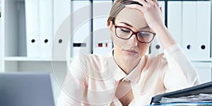 Tired and exhausted woman in spectacles looks down near the mountain of documents propping up her head with her hands.