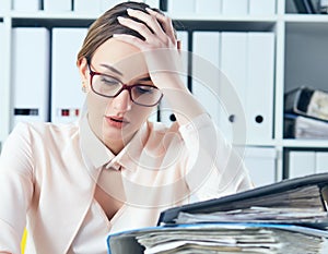 Tired and exhausted woman in spectacles looks down near the mountain of documents propping up her head with her hands.