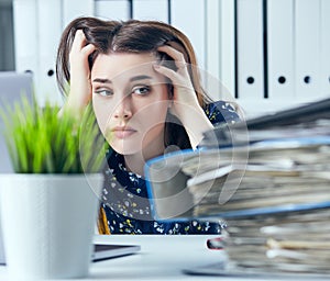 Tired and exhausted woman in spectacles looks at the mountain of documents propping up her head with her hands.