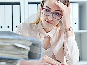 Tired and exhausted woman in spectacles looks at the mountain of documents propping up her head with her hands.