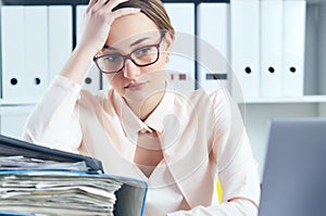 Tired and exhausted woman in spectacles looks at the mountain of documents propping up her head with her hands.