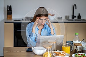 Tired exhausted workaholic woman with headache working on laptop sitting at table in kitchen.