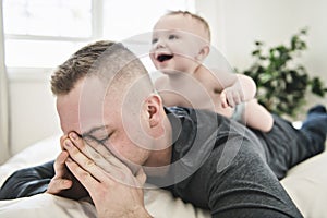 Tired father sleep but his baby boy interferes him. Young happy father lying on bed while his son play with him