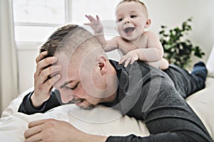 Tired father sleep but his baby boy interferes him. Young happy father lying on bed while his son play with him