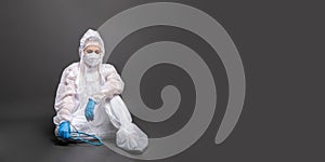 Tired female doctor in a protective suit and mask sitting on the floor on a gray background. Banner image with copy