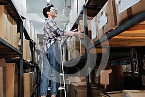 Tired female warehouse worker standing on the ladder, placing box on the shelf