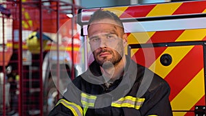 Tired fireman in protective uniform looking at camera while sitting near fire engine on station