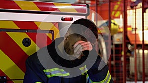 Tired fireman in protective uniform looking at camera while sitting near fire engine on station