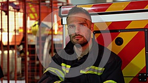 Tired fireman in protective uniform looking at camera while sitting near fire engine on station after extinguishing a