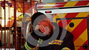 Tired fireman in protective uniform looking at camera while sitting near fire engine on station after extinguishing a