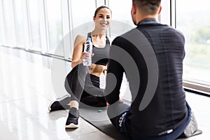 Tired fitness couple having rest and talking, drinking water at gym. Man and woman sitting on floor, break after training