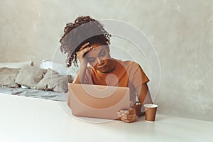 Tired flustrated African descent young girl sitting at desk in front of laptop while irritably looking at computer screen with