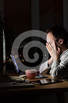 Tired freelancer male rubbing his eyes, sitting at a desktop PC/laptop late at night, falls asleep from fatigue