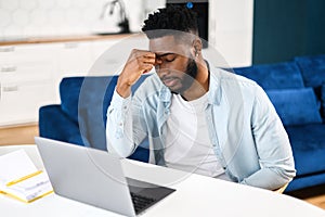Tired frustrated black man sitting with eyes closed at the table and using laptop at home