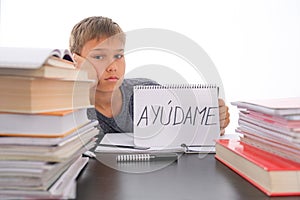 Tired frustrated boy sitting at the table with many books, exercises books. Spanish word Auydame - Help me- is written photo