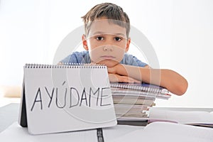 Tired frustrated boy sitting at the table with many books, exercises books. Spanish word Auydame - Help me- is written photo