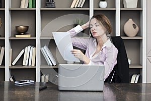 Tired frustrated and overworked business woman sitting in the office, holding documents and reading about problems in business