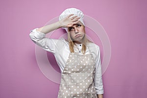 Tired girl cook in apron covers her face with a hand on a pink background, sad housewife in kitchen clothes in stress