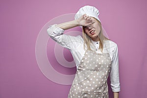 Tired girl cook in apron covers her face with a hand on a pink background, sad housewife in kitchen clothes in stress