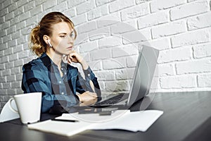 Tired girl freelancer sitting at table with laptop on background of white brick wall