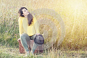 Tired girl with hat sitting and resting on brown vintage suitcase in the field road during summer sunset. Toned image and travel c