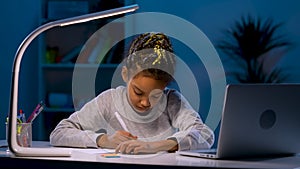 Tired girl reluctantly rewrites the task from the laptop. An African American girl sits at a table in a dark room by the