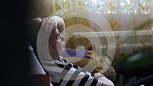 A tired girl sits on the sofa in the living room after a hard day and puts her hand to her head trying to relax