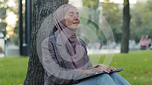 Tired girl student in hijab young business woman with laptop sitting in park near tree on green lawn turns off closes