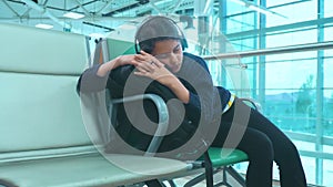 Tired girl teen in headphones traveler sleeping on the airport waiting for the plane departure gates bench with